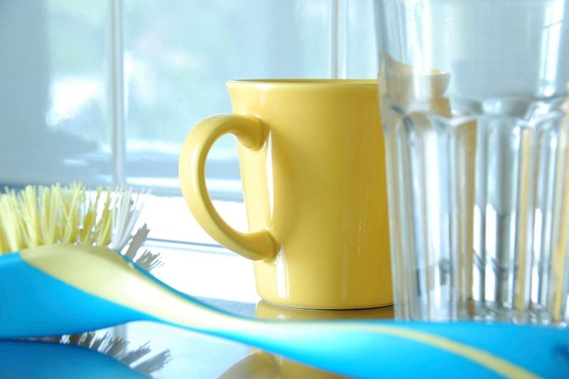 Cepillo de fregar, taza amarilla y vaso secándose junto a una ventana luminosa