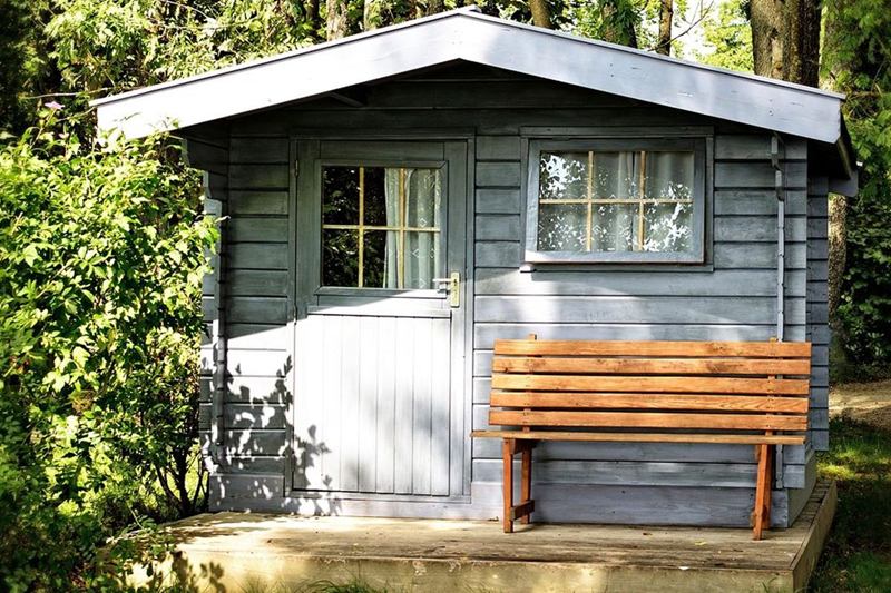 Caseta de jardín de madera gris con ventanas y un banco delante