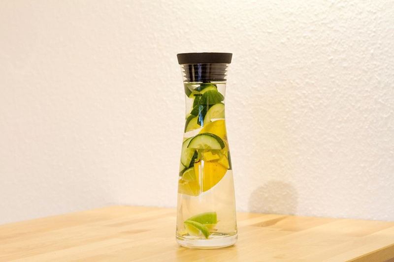 Jarra de cristal con agua, limón y pepino sobre una mesa de madera clara