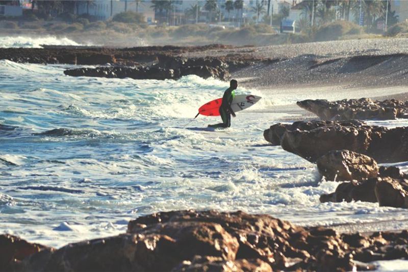 Surfista con traje de neopreno y tabla roja saliendo del mar entre las rocas